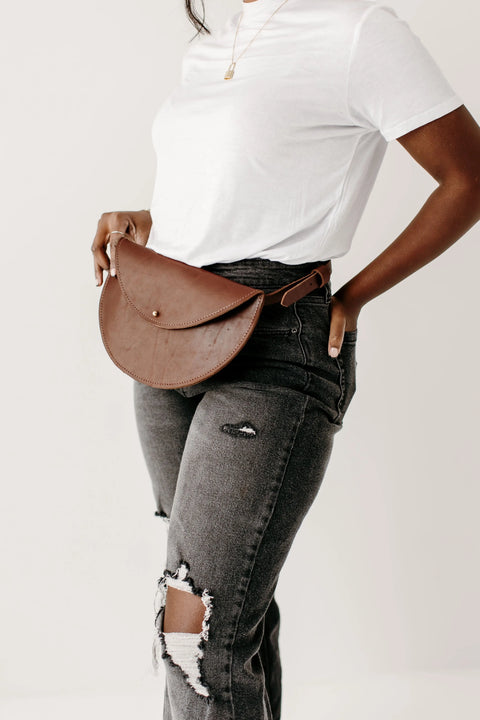 A woman wearing a white t-shirt and dark ripped jeans stands against a white background, accessorized with the Urban Southern Half-Moon Belt Bag—a brown leather fanny pack handcrafted in the USA—around her waist.