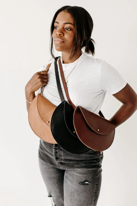 A woman in a white t-shirt and jeans models three Urban Southern Half-Moon Belt Bags, each made from full-grain leather in the USA, draped crossbody over her shoulder in front of a plain white background.