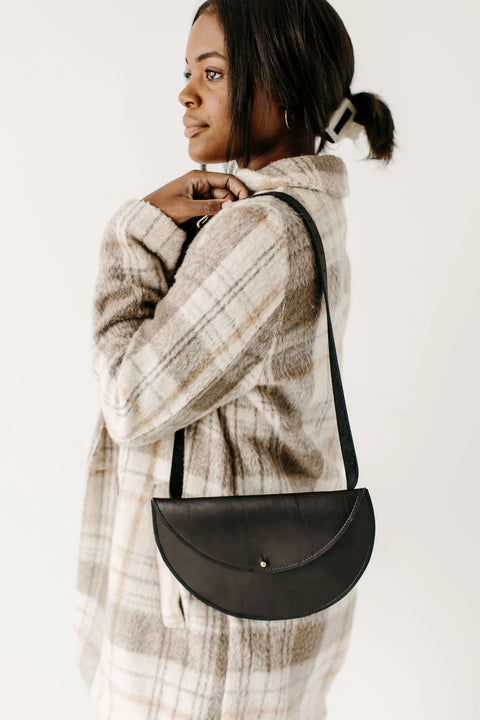 A woman in a plaid coat stands sideways, wearing the Urban Southern Half-Moon Belt Bag—handcrafted in the USA—over her shoulder. Her dark hair is pulled back with a clip as she looks forward against a plain light background.