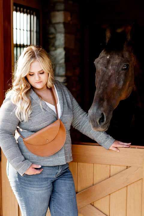 A woman with long blonde hair, wearing a gray jacket, jeans, and the Urban Southern Half-Moon Belt Bag, stands beside a dark brown horse, resting her hand on a wooden stall door inside a stable.