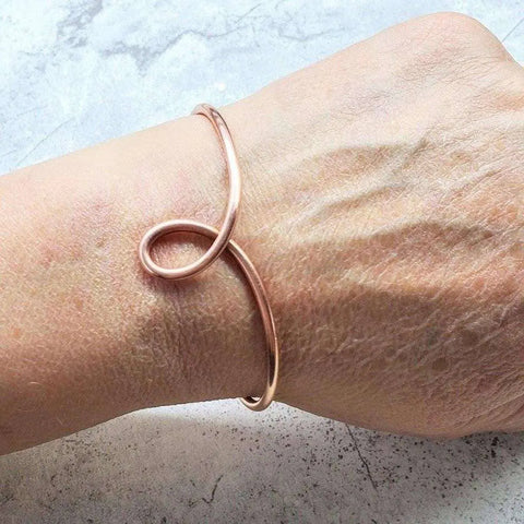 A hand wears the Handmade Copper Teardrop Bangle by Alexa Martha Designs, a minimalist, handcrafted bracelet featuring an elegant loop shape against a light, textured background—an adjustable piece exuding refined sophistication.