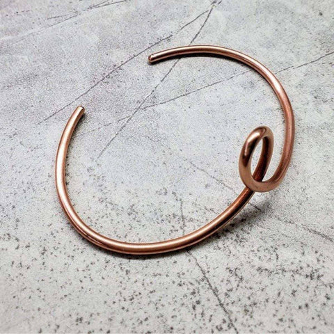 The Handmade Copper Teardrop Bangle by Alexa Martha Designs features a minimalist open-ended design with a smooth loop at one end, displayed on a textured gray surface—a striking piece of handcrafted jewelry.