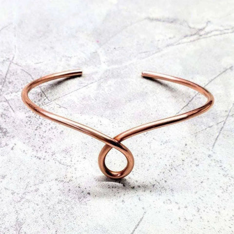 The Handmade Copper Teardrop Bangle by Alexa Martha Designs is a minimalist adjustable bracelet with a teardrop design, crafted from polished copper and showcased on a light, textured stone surface.