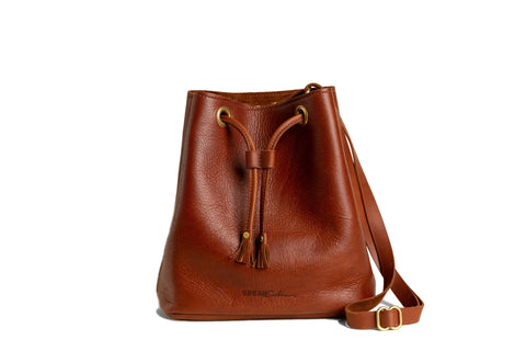 The Urban Southern Urban Bucket Bag is a brown leather bag handcrafted in the USA, featuring a drawstring closure, tassel accents, and an adjustable shoulder strap, shown against a white background.