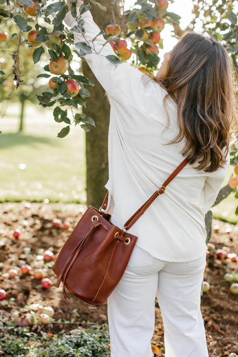 A woman with long brown hair, in a white shirt and pants, stands outdoors picking an apple from a tree. She carries the Urban Southern Urban Bucket Bag over her shoulder, with apples scattered on the ground.