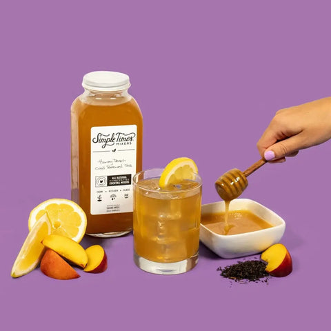 A bottle of Simple Times Mixers Honey Peach Cold Brewed Tea, a glass of iced tea with lemon and peach slices, organic black tea leaves, and honey—all set on a purple background. Perfect as a cocktail mixer!.
