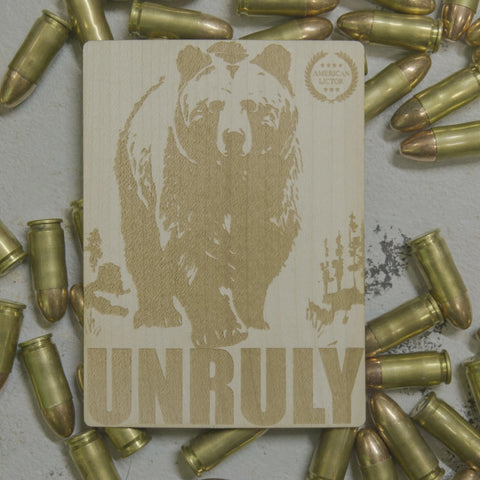 A Wood Sticker by American Lictor, featuring a bear and UNRULY engraved on wood, with scattered bullets atop a gray surface. The plaque also displays the American Lictor emblem and resembles laser-etched wood stickers.
