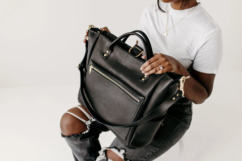 A woman in a white tee and ripped black jeans holds Urban Southern’s Legacy Backpack Tote, featuring gold hardware, convertible straps, and a front zipper pocket, while seated against a white background.