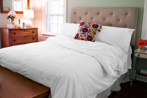 A cozy bedroom featuring the American Blossom Linens Cotton Duvet Cover Set, accented by a colorful floral pillow, tufted headboard, wooden dresser, flowers, lamp, and natural light showcases quality craftsmanship.