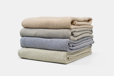 Four Herringbone Weave Cotton Blankets by American Blossom Linens in beige, gray, blue, and cream are neatly stacked, bringing a cozy touch to any room against a simple white background.