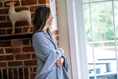 A woman wrapped in the Herringbone Weave Cotton Blanket by American Blossom Linens stands by a large window, next to a brick wall with a shelf holding a decorative fabric deer.