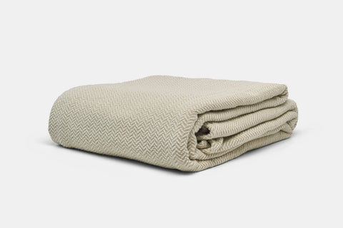 The Herringbone Weave Cotton Blanket by American Blossom Linens, shown in beige on a white background, features a textured herringbone design that adds cozy comfort and timeless charm to any room.