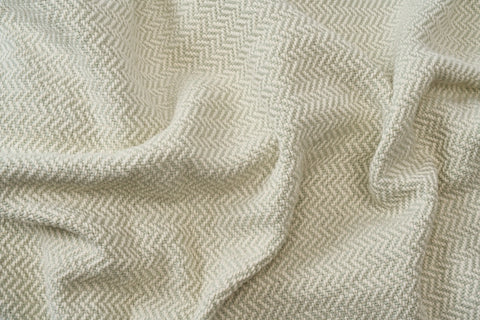 A close-up of the Herringbone Weave Cotton Blanket by American Blossom Linens shows its light-colored, textured fabric with a subtle herringbone pattern softly draped in gentle folds and shadows.
