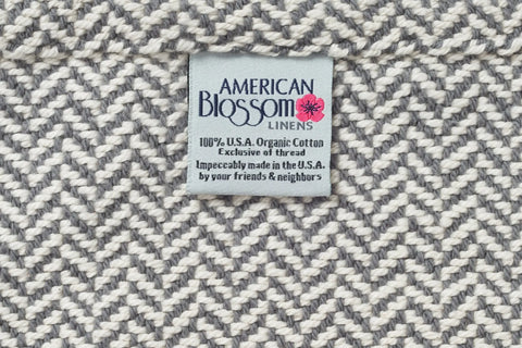 The fabric label reads: American Blossom Linens. Herringbone Weave Cotton Blanket. 100% U.S.A. Organic Cotton, exclusive of thread. Impeccably made in the U.S.A., this heirloom-quality blanket features a textured gray and white woven pattern.