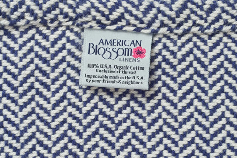 A fabric tag on the Herringbone Weave Cotton Blanket by American Blossom Linens reads, “100% U.S.A. organic cotton, exclusive of thread, impeccably made in the U.S.A. by your friends & neighbors.”.