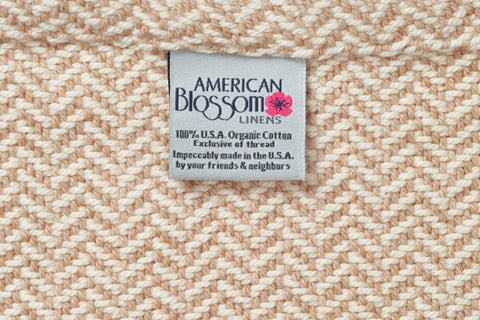 A close-up of the Herringbone Weave Cotton Blanket with a label reading American Blossom Linens. Made from 100% U.S.A. organic cotton, this heirloom-quality blanket is expertly crafted in the U.S.A.