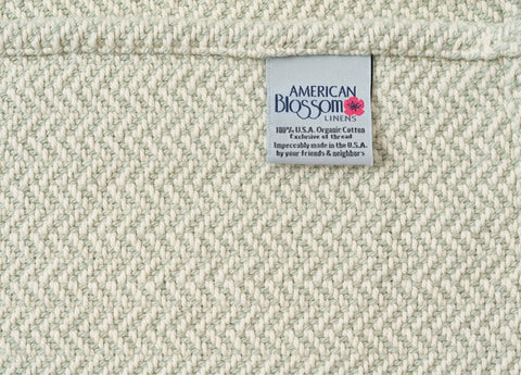 A close-up of the Herringbone Weave Cotton Blanket by American Blossom Linens shows a tag reading “100% U.S.A. Organic Cotton, Impeccably made in the U.S.A.”—an heirloom-quality, cream-colored woven layer for any home.