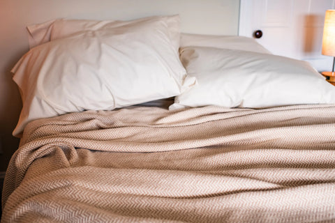 A neatly made bed with two large white pillows and an American Blossom Linens Herringbone Weave Cotton Blanket, softly illuminated by warm lamp light, adds a cozy touch to the inviting scene.