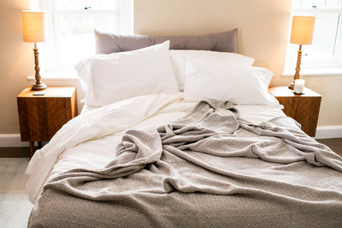 A cozy, unmade bed with white sheets, pillows, and an American Blossom Linens Herringbone Weave Cotton Blanket sits between two wooden nightstands with lamps in a softly lit bedroom featuring two windows.