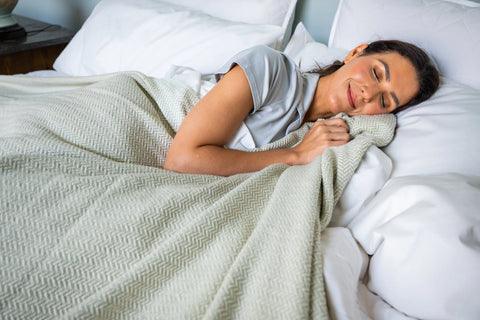A dark-haired woman smiles peacefully while sleeping on her side, wrapped in the American Blossom Linens Herringbone Weave Cotton Blanket, adding a perfect cozy layer to her white bed.