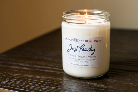A lit Hand Poured Soy Wax Scented Candle by American Blossom Linens rests on a wooden surface, its label reading “Just Peachy - Citrus + Peach + Vanilla,” and filling the air with a delightful local fragrance.