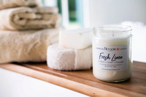 The Hand Poured Soy Wax Scented Candle by American Blossom Linens, labeled “Fresh Linen,” sits on a wooden surface beside beige towels and a white soap bar, creating a clean, cozy spa-like atmosphere with its refreshing scent.