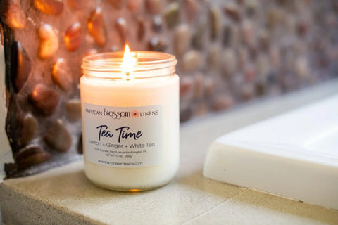A Hand Poured Soy Wax Scented Candle by American Blossom Linens labeled “Tea Time, Lemon + Ginger + White Tea” sits on a bathroom counter near a white tub and stone-tiled wall, filling the space with its amazing fragrance.