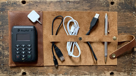 The Rogue Industries Leather Tech Organizer holds a PayPal card reader, white card reader adapter, two USB charging cables with cord holders, black stylus, and silver pen—all neatly arranged on a wooden surface.