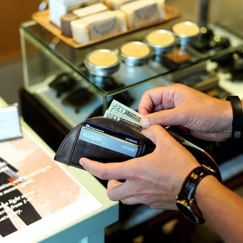 Someone holds a Rogue Industries Rogue Front Pocket Wallet in Ballglove open with cash and a bank card, ready to pay at a counter displaying soaps and jars in the background.