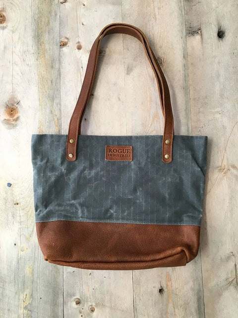 The Saco River Tote Bag by Rogue Industries features full-grain leather straps and base, a gray fabric upper, and a branded leather patch. Crafted in Maine, its showcased on a rustic wooden surface.