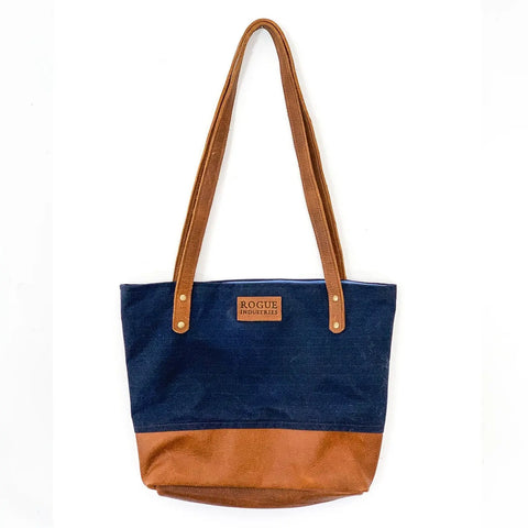 The Saco River Tote Bag by Rogue Industries features a two-tone waxed canvas design with a navy blue upper, brown base, full-grain leather straps, and a front center Rogue Industries logo patch. Shown on a plain white background.