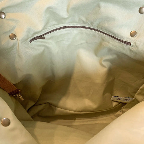 The image shows the interior of the Rogue Industries Saco River Tote Bag, featuring light-colored waxed canvas, a brown zipper pocket, metal snap buttons along the edges, a full-grain leather strap, and a small side fabric tag.