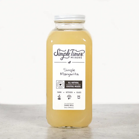 A clear glass bottle of Simple Times Mixers’ Simple Margarita, filled with light yellow liquid and made with organic cane sugar, sits on a gray surface against a white background.