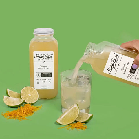 A bottle of Simple Times Mixers Simple Margarita is poured over ice in a glass, surrounded by fresh lime wedges and blood orange zest on a green background, with another Simple Margarita bottle standing upright nearby.