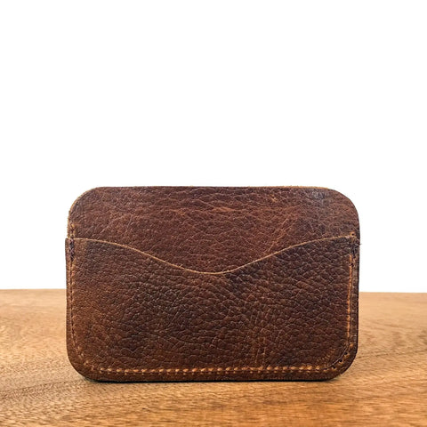 The Rogue Industries Portland Card Case, crafted in Maine from genuine brown leather with visible stitching and a textured finish, rests on a wooden surface against a plain white background.