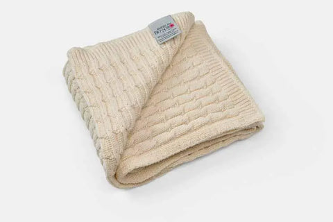 The American Blossom Linens Cotton Knitted Baby Blanket, in cream with a textured cable pattern, is neatly folded on a white background. A label with text is attached to one corner—ideal for crib or stroller use.