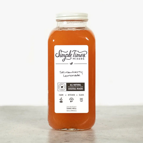 A clear glass bottle of Simple Times Mixers Strawberry Lemonade cocktail mixer, filled with orange-pink liquid, is placed on a light surface against a plain white background.