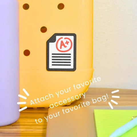 Chatelet Manufacturing FRESHe Baglets Teacher Charms decorate your tote bag with style! The yellow charm has round holes and an A+ paper patch—attach to Bogg Bags, Simply Southern, or any teacher tote. Purple bottle and green notepad shown nearby.