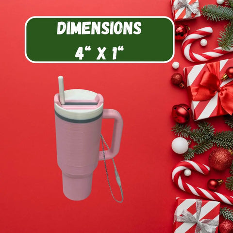 The Chatelet Manufacturing Tumbler Cup Replica Christmas Ornament, styled like a pink-handled tumbler with straw and strap, is shown on a red background amid holiday décor, gifts, and tree ornaments. A green sign above reads DIMENSIONS 4” x 1” in white text.