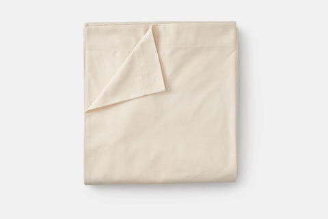 A neatly folded beige Cotton Single Flat Bed Sheet by American Blossom Linens is displayed on a white background, with one corner turned over to show its inner side—crafted from luxurious 100% USA grown cotton.