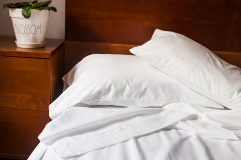 A neatly made bed with a luxurious American Blossom Linens Cotton Single [Separate] Flat Bed Sheet sits beside a wooden nightstand holding a potted plant in a white pot labeled BLOSSOM.