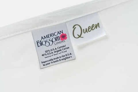 Close-up of fabric tags on white bedding, showing American Blossom Linens Cotton Single [Separate] Flat Bed Sheet, Queen size, crafted from luxurious 55% U.S.A. cotton and 45% organic cotton with Impeccably made in the U.S.A. noted.