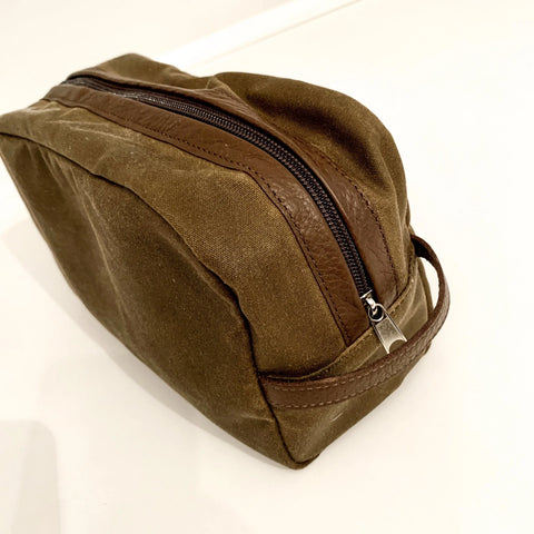 The Rogue Industries Waxed Canvas Dopp Kit, featuring leather accents, a sturdy zipper, and side handle, is shown on a white surface—durable and convenient for travel essentials.