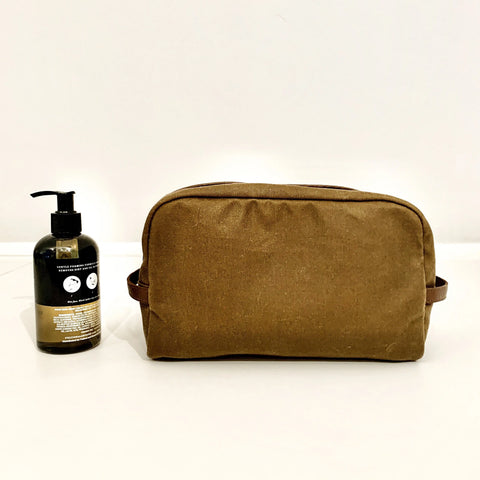 The Rogue Industries Waxed Canvas Dopp Kit in brown, featuring side handles, sits on a white surface beside a dark pump bottle with a visible label, against a plain white background. This stylish shave kit is made in the USA.