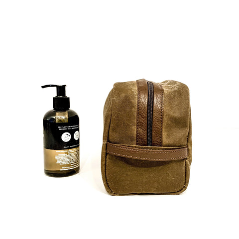 A Rogue Industries Waxed Canvas Dopp Kit in brown stands upright beside a black pump bottle, both displayed on a plain white background.