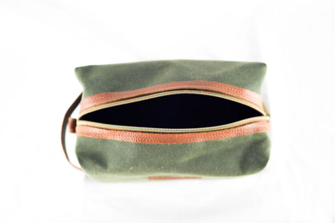 The Rogue Industries Waxed Canvas Dopp Kit, in green with a brown leather zipper, is shown from above with its zipper partially open to reveal the empty interior. This stylish shave kit is made in the USA and set against a white background.