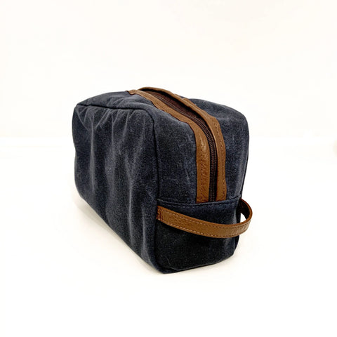 The Rogue Industries Waxed Canvas Dopp Kit, a rectangular dark gray bag with a brown leather zipper and handle, made in the USA, is shown on a plain white surface.