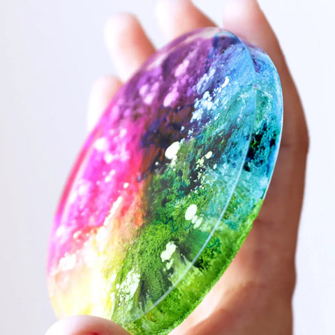 A hand holds a round resin disc with vibrant colors blending from pink and purple to green and blue, accented by white flecks made with ArtResin Alcohol Ink Sinker for a marbled, abstract petri art coaster design.