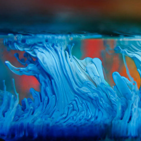 ArtResin’s Alcohol Ink Sinker creates abstract, close-up swirls of blue and white ink under water, forming wave-like patterns against a blurred background with warm orange and red tones.